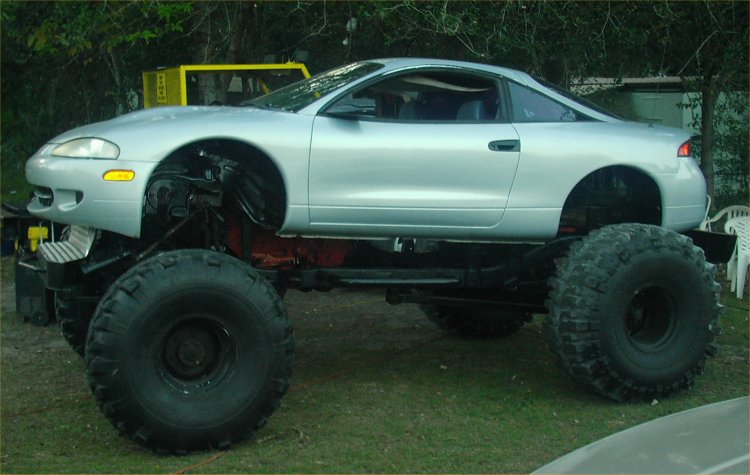 Silver coupe body on a massive off-road chassis with huge tires. Yellow vehicle in background