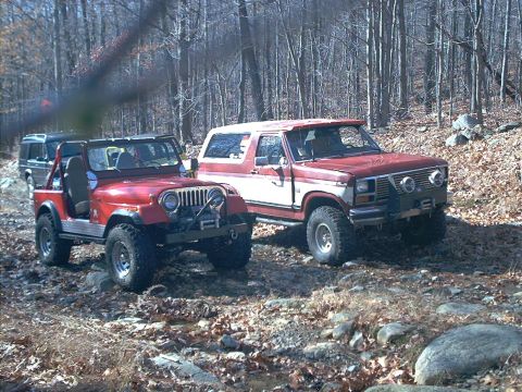1986 Ford Bronco - Two off-road vehicles, a Ford Bronco and a Jeep, are parked on a trail surrounded by trees.