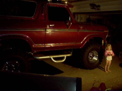 1978 Ford Bronco - A red 1978 Ford Bronco is shown in profile, a lifted suspension and side steps, with a small child standing nearby.