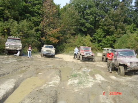 1978 Ford Bronco - Several off-road vehicles navigate a muddy track in a wooded area, with people standing nearby.