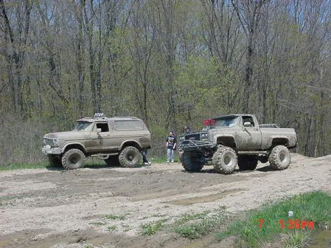 1978 Ford Bronco - Two modified off-road vehicles are parked on a dirt path surrounded by trees.