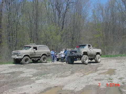 1978 Ford Bronco - Two heavily modified trucks are parked on a muddy trail surrounded by trees, with a group of people observing.
