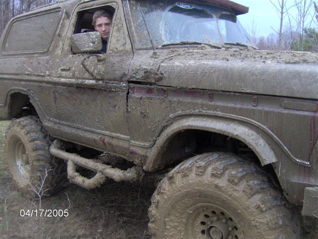Mud-covered SUV with a person in the driver's seat, large off-road tires, and a cracked fender
