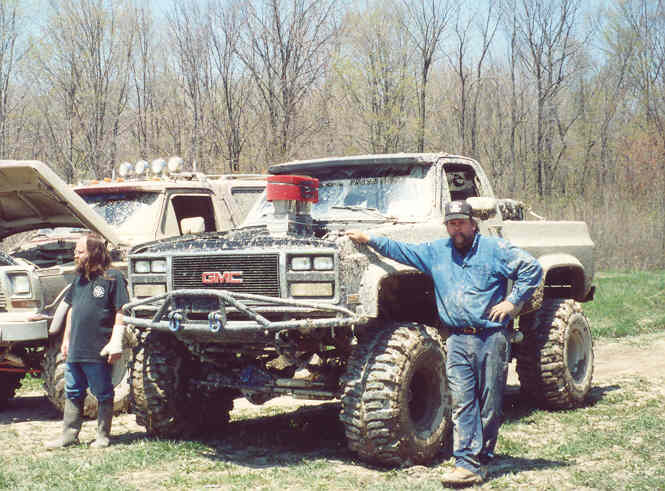 Mud-covered GMC pickup truck with large tires and exposed engine, a man leans on it
