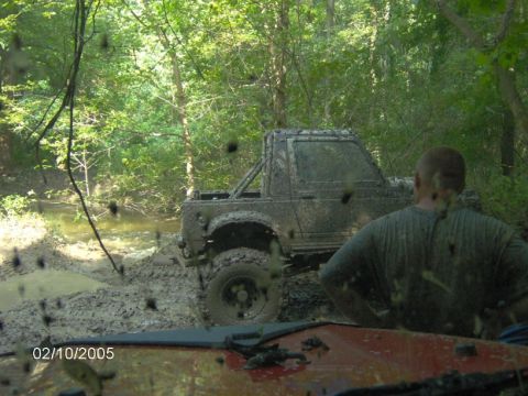 1978 Ford Bronco - A muddy off-road vehicle navigates a wooded stream, with another vehicle visible in the foreground.