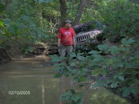 1978 Ford Bronco - Man stands in muddy water next to a stuck Jeep partially submerged in a wooded area.