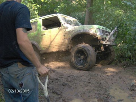 1978 Ford Bronco - A person with a shovel stands next to a mud-splattered off-road vehicle attempting to navigate a muddy trail.