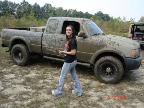 1978 Ford Bronco - A mud-covered extended-cab pickup truck parked in an off-road field, with a young woman in a black t-shirt and jeans walking past it. The t…