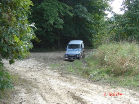 1978 Ford Bronco - A blue SUV is stuck in the mud on a dirt road surrounded by trees and tall grass.