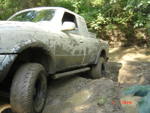 1978 Ford Bronco - This pickup truck is covered in mud as it drives through a challenging muddy trail surrounded by trees.