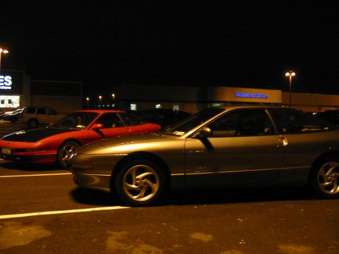 1996 Ford Probe - Two Ford Probes, a silver one and a red one with a black hood, are parked in a lot after dark.