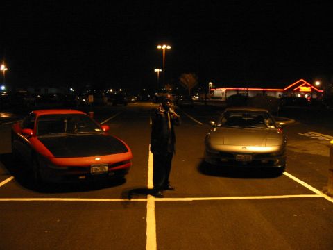 1996 Ford Probe - Two sports cars, one red and one silver, are parked in a lot at night with a person standing between them.