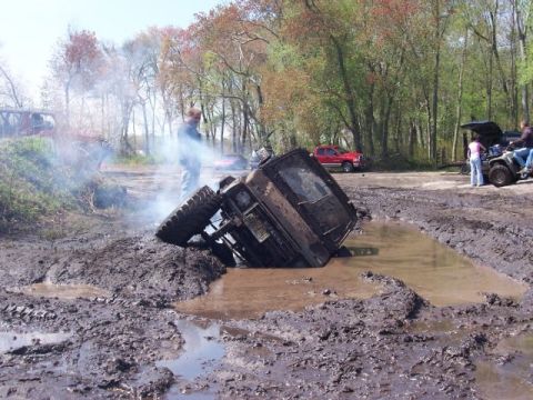1987 Suzuki Samurai - An off-road vehicle is stuck in a deep mud pit while other vehicles and people are visible in the background.