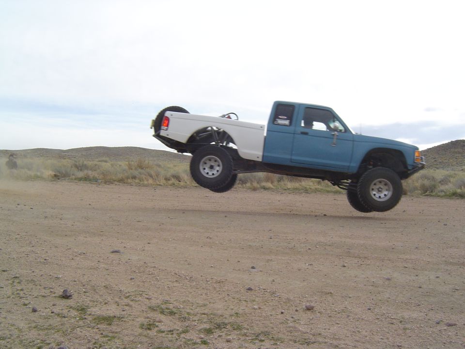 Blue and white pickup truck mid-air over a dirt road, with a spare tire mounted on the back