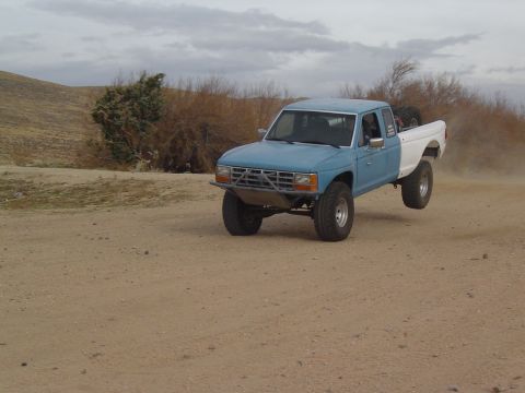 1985 Ford Ranger - A modified pickup truck jumps over a dirt track, kicking up dust in a dry, barren landscape.