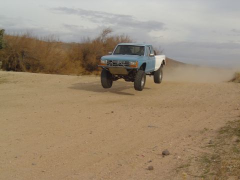 1985 Ford Ranger - A blue and white 1985 Ford Ranger is captured mid-air during a jump on a dusty dirt road.
