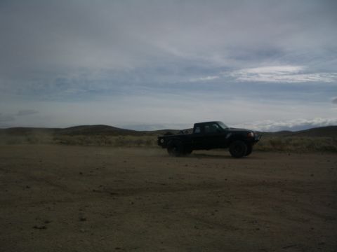 1985 Ford Ranger - A black pickup truck drives on a dusty dirt road under a cloudy sky.