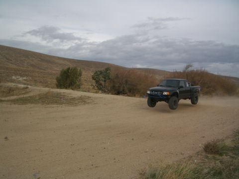 1985 Ford Ranger - A black pickup truck is captured airborne while driving on a dusty, unpaved road with rolling hills in the background.