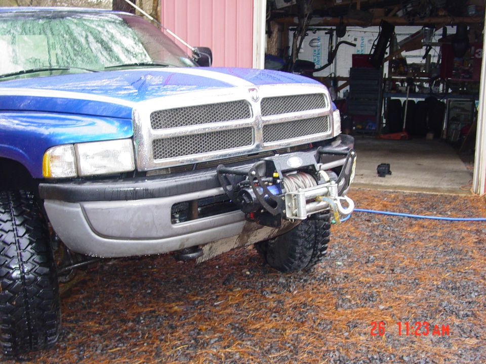 Front view of a blue Dodge pickup truck with a winch mounted on the bumper. The truck has a chrome grille and white racing stripes on the hood