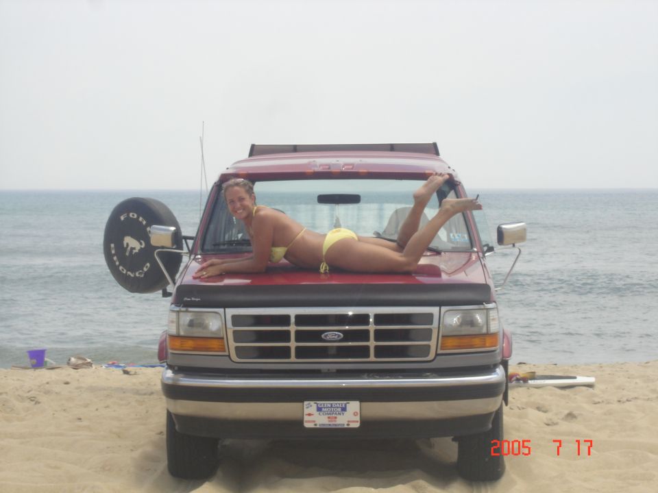 Red Ford Bronco parked on beach, front view with woman posing on hood