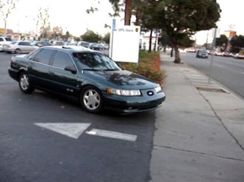 1993 Ford Taurus - A dark green 1993 Ford Taurus is parked on the side of a road next to a sidewalk.