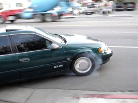 1993 Ford Taurus - Green 1993 Ford Taurus performing a burnout on a city street with traffic in the background.