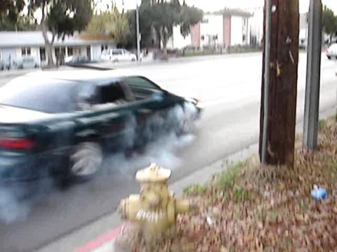 1993 Ford Taurus - A dark green sedan is seen doing a burnout on a street, with smoke billowing from its tires.