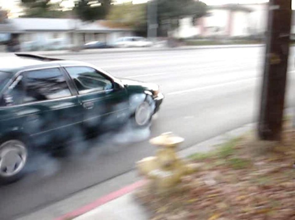 Dark green sedan doing a smoky front-wheel burnout on a street by a yellow fire hydrant