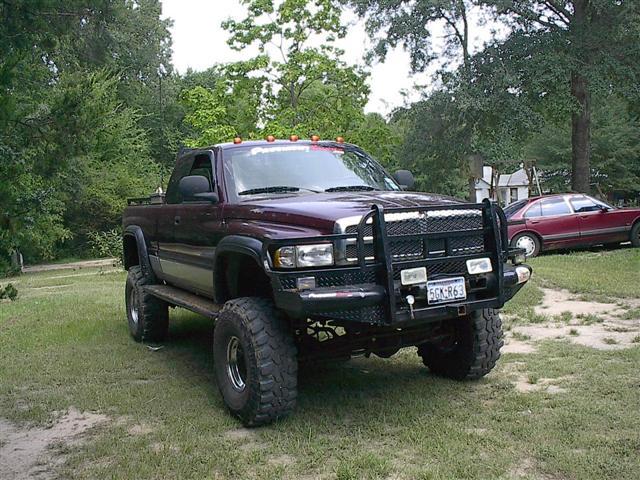 Lifted maroon Dodge pickup truck with bull bar and aftermarket wheels, parked on grass. Red sedan in background