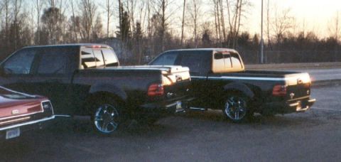 2003 Ford F-150 - Two black pickup trucks are parked next to each other on a paved surface with trees in the background.