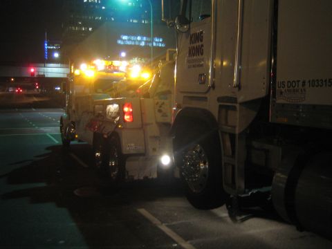 1968 Ford Mustang - A tow truck is positioned behind a semi-trailer truck, both illuminated by the tow truck's flashing lights on a city street at night.