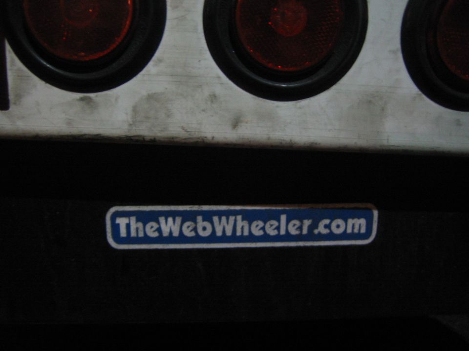 Close-up of dirty white panel with four red tail lights above a blue sticker reading "TheWebWheeler.com"