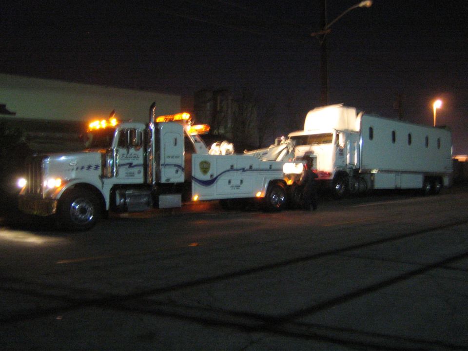 White tow truck with amber lights is towing a white trailer at night. Text on tow truck reads 'PACE'