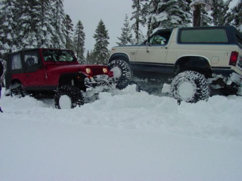 1986 Ford Bronco - Two off-road vehicles struggle in a snowy forest, with the Bronco appearing to be from the early 1980s.