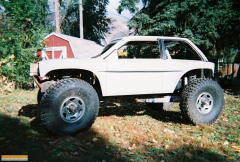 1991 Ford Bronco - A modified white vehicle with oversized tires sits in a grassy area next to a red shed.