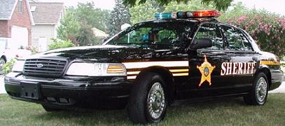 2000 Ford Crown Victoria - A black sheriff's car is parked on a grassy area with houses and trees in the background.
