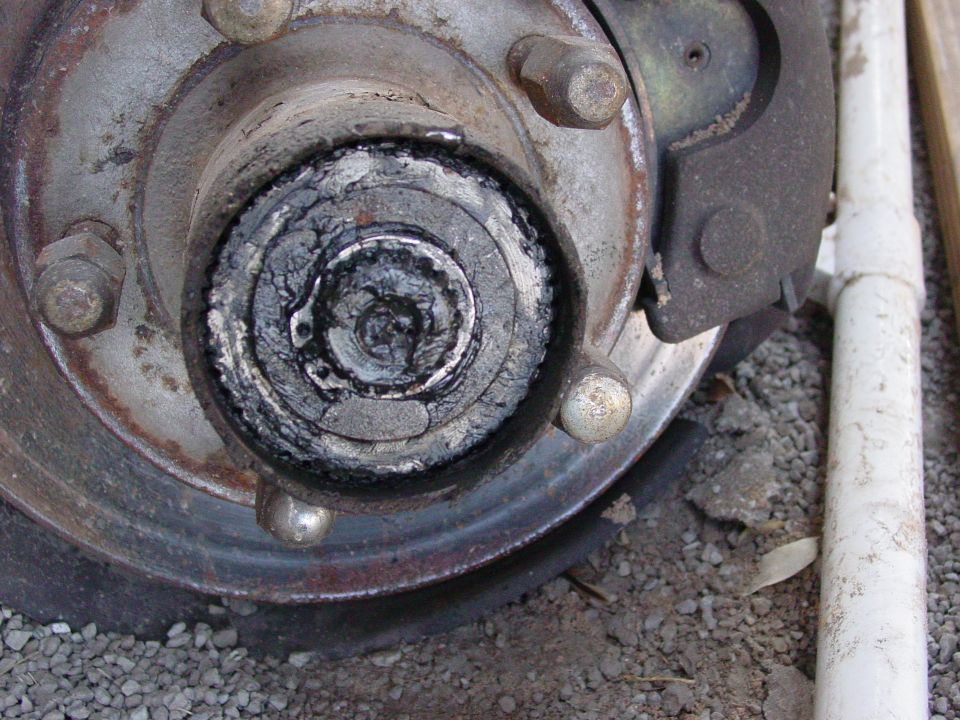 Close-up of greasy wheel hub with cap removed showing spindle nut, lug studs, and rusty drum on gravel