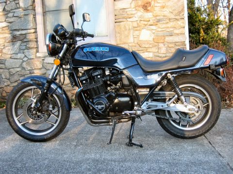 1985 Ford LTD Crown Victoria - Left-side profile of a dark blue Suzuki GS1100E standard/naked motorcycle on its center stand, showing the air-cooled inline-four engine, c…