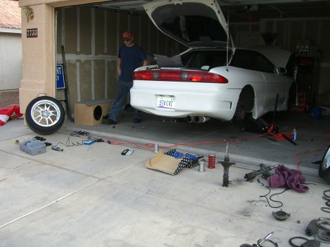 1996 Ford Probe - A white coupe is parked in a garage with its trunk open and various car parts scattered on the floor, suggesting maintenance or repair work.