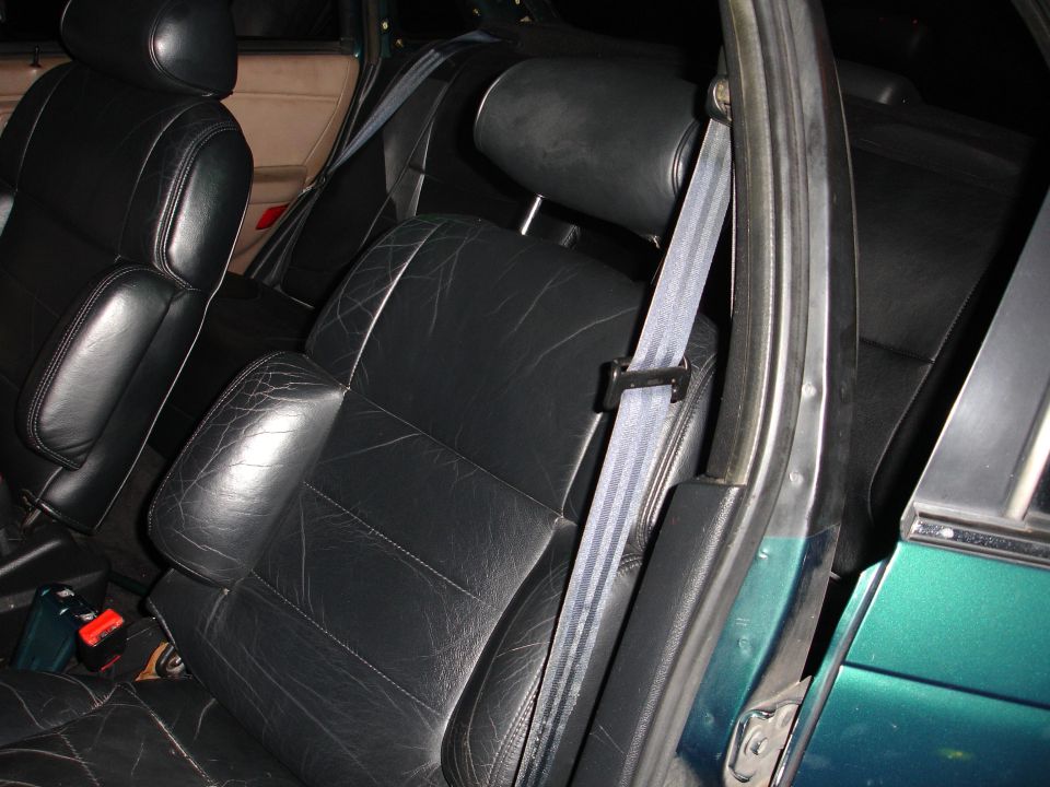 Interior view of car seats, showing black leather with visible wear and stitching. Seatbelts are buckled