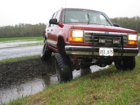 1991 Ford Explorer - A red 1991 Ford Explorer is shown with a lifted suspension and off-road tires, navigating through a muddy roadside puddle.
