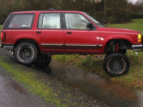 1991 Ford Explorer - A red 1991 Ford Explorer SUV is shown with a lifted suspension, navigating through a muddy, watery ditch.