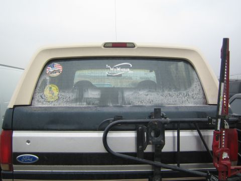 1990 Ford Bronco - Rear view of a dark Ford SUV with a fogged rear window, a Web Wheeler windshield banner, US flag sticker, and a red Hi-Lift jack on the bum…