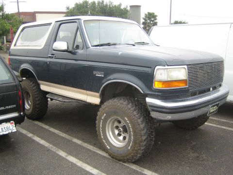 1990 Ford Bronco - dark green lifted Ford Bronco on chrome wheels and mud tires sits in a parking lot beside a black Chevy Blazer and a white van.
