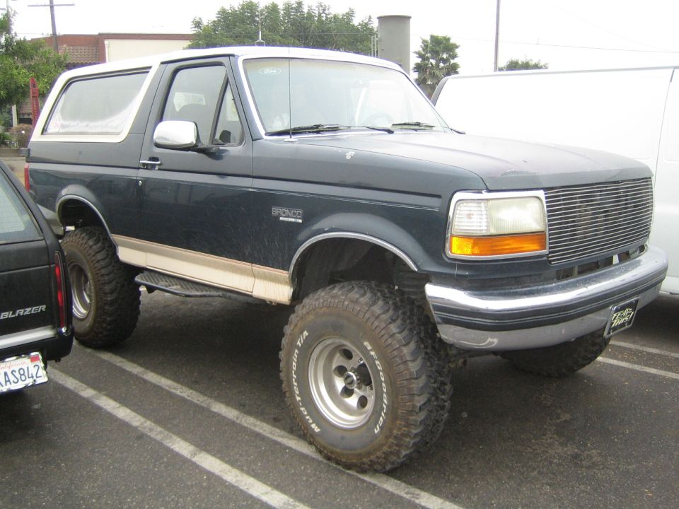 Dark green lifted Ford Bronco on mud tires parked in a lot next to a Chevy Blazer