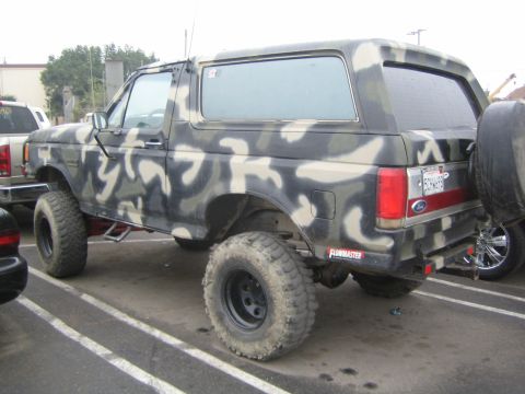 1990 Ford Bronco - Rear three-quarter view of a camouflage-painted lifted Ford Bronco on black steel wheels and mud tires, parked in a paved lot.