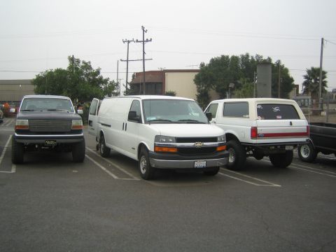 1990 Ford Bronco - Three vehicles parked side by side in a lot: a dark lifted Ford, a white Chevrolet Express van, and a white lifted Ford SUV.