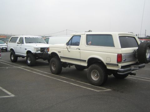 1990 Ford Bronco - Two white lifted Ford Broncos on mud tires parked at angles in a paved lot under an overcast sky, one with a rear-mounted spare tire.
