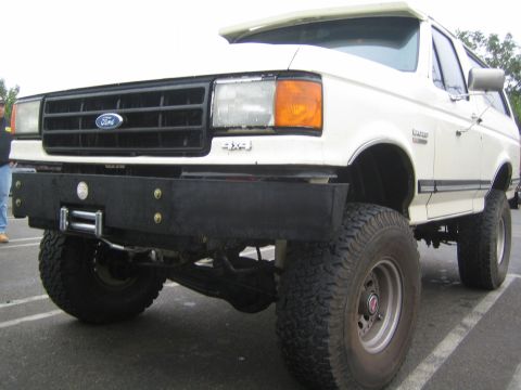 1990 Ford Bronco - Front three-quarter view of a white lifted Ford Bronco with a black winch bumper, Ford grille, 4x4 badge and mud tires on steel wheels.