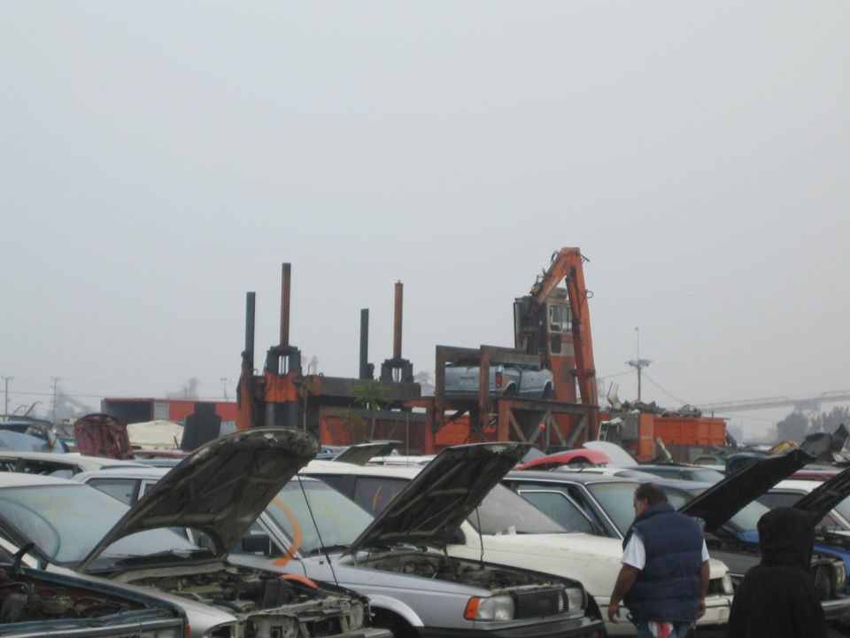 Salvage yard with rows of cars with open hoods and an orange crusher or loader machine behind